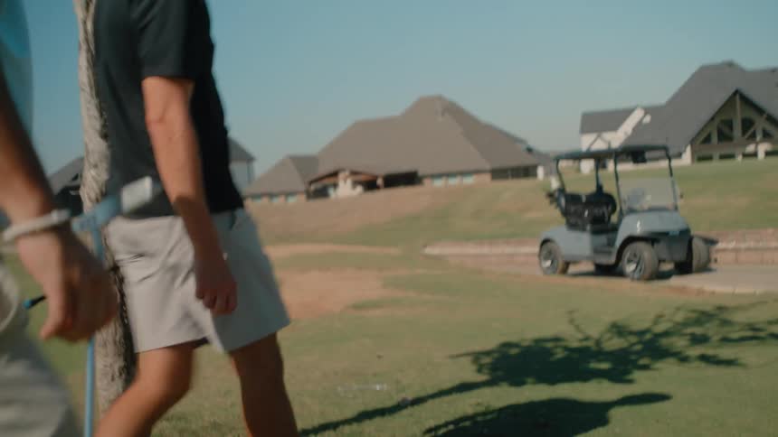 The video shows two men walking across a sunny golf course toward a parked golf cart in the background several large suburban houses line the edge of the green under a clear blue sky