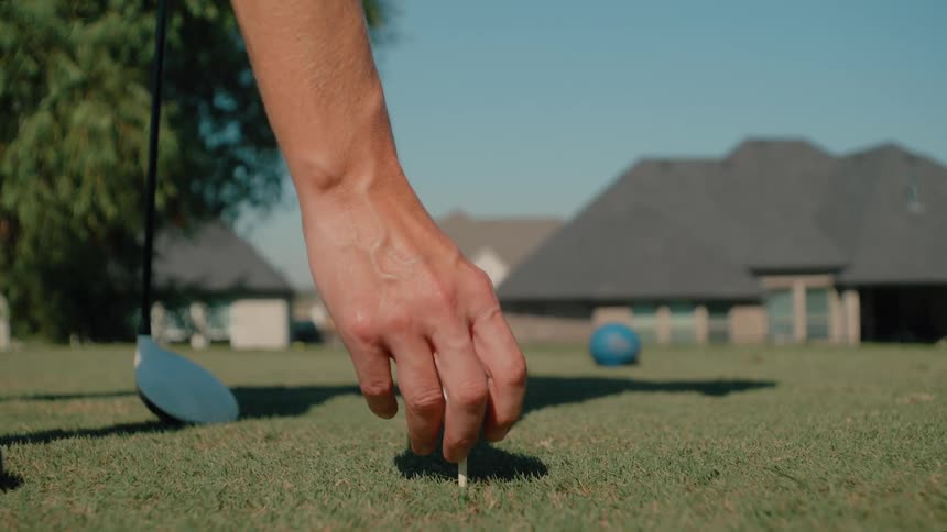 A mans hand places a white golf ball onto a wooden tee in the grass while a driver rests on the ground nearby with suburban houses and a blue lawn ornament blurred in the sunny background