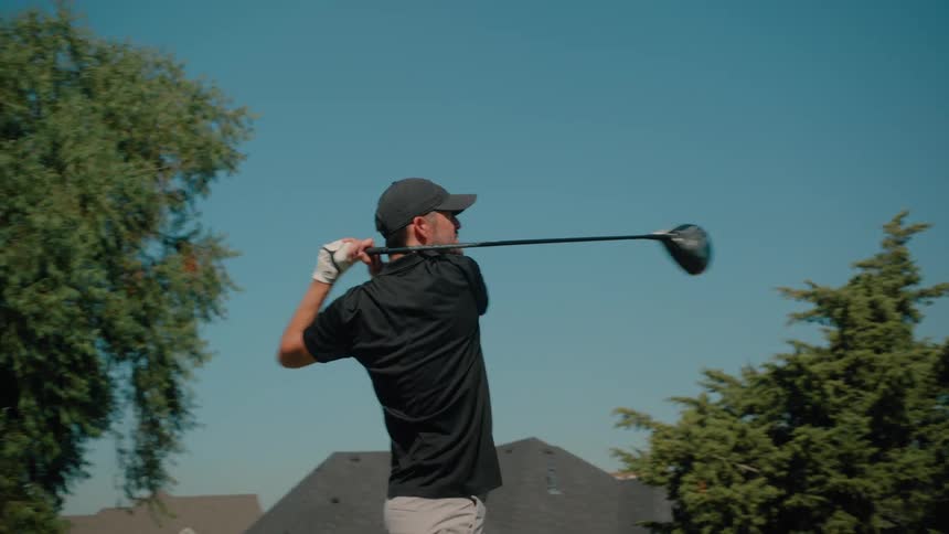 A man in a black polo and grey shorts performs a full golf swing with a driver on a grass range then he follows the balls flight against a backdrop of suburban houses and green trees under a clear blue sky during a sunny day