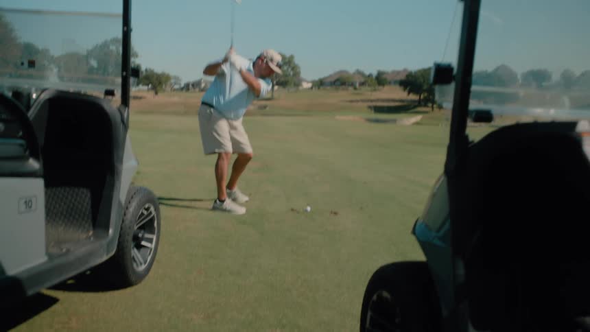 Male golf player hitting a ball with a golf club next to two carts outdoors during day time