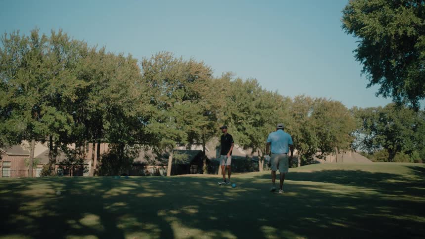 Two men walk across the field with their golf clubs as one man sets a golf ball on the grass