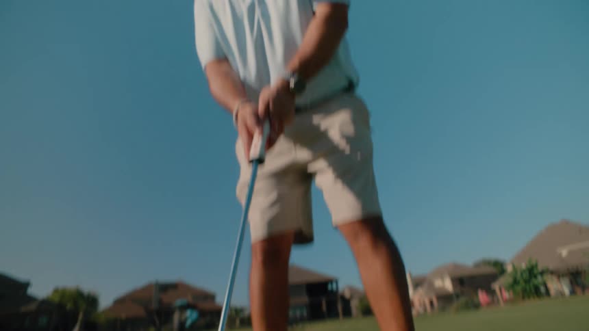 A man in a light blue polo shirt and tan shorts practices his putting on a sunny golf course the camera uses a lowangle shot to capture the movement of the putter hitting the ball toward the hole