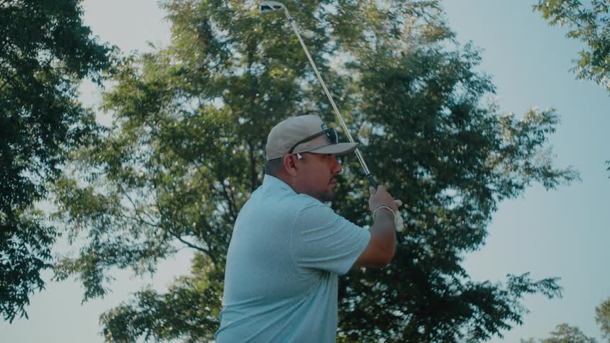 A man in a light blue polo and tan cap performs a golf swing on a sunny day and he strikes the ball and watches its flight against a background of lush green trees and a clear sky before walking forward