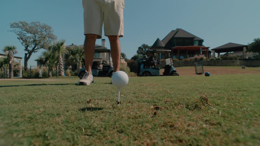Male golf player placing a ball and hitting it with a golf club with golf carts and another man on the background outdoors during day time