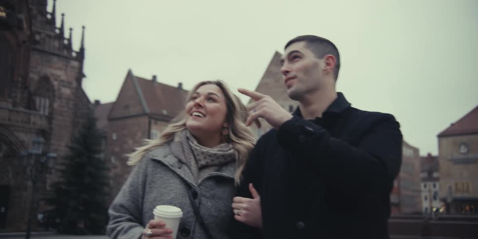 A happy couple strolls arminarm through an old european town square smiling and chatting while sharing a coffee on an overcast day