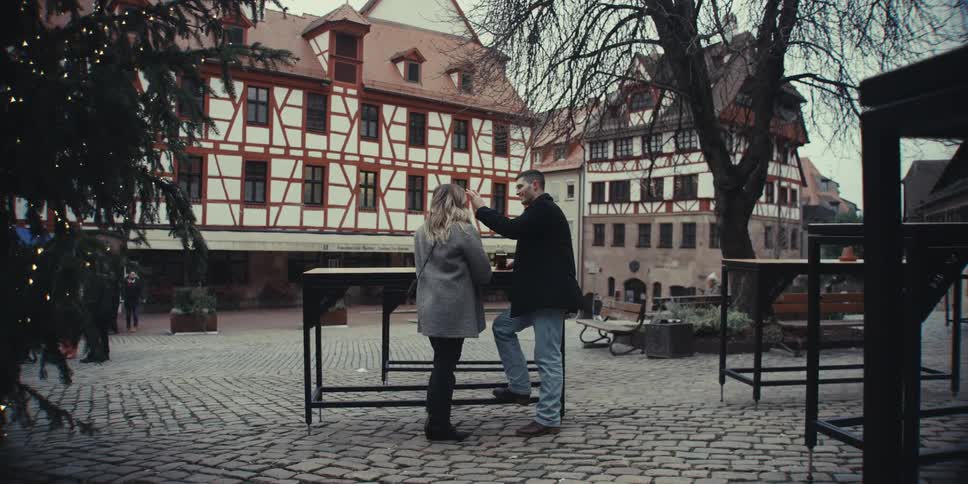 A young couple standing outdoors at midday talking beside a tall table with buildings rising in the background