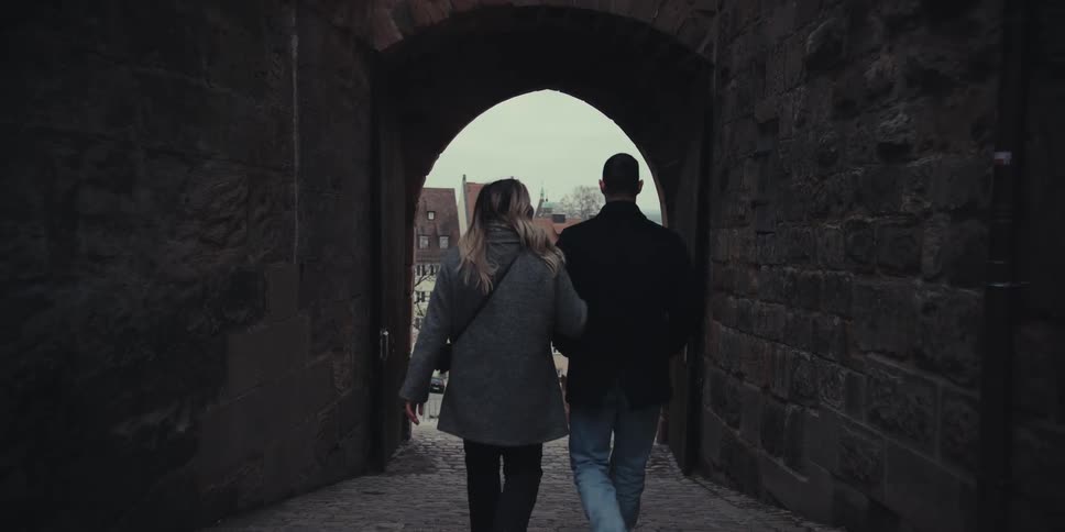 A couple holding hands walks out of a tunnel into the street at midday stepping from the shaded passage into bright sunlight