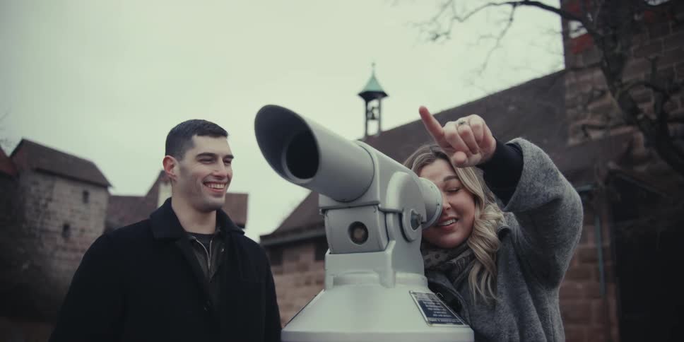 A woman points excitedly while looking through a public viewing telescope as a man stands beside her before they walk away at a historic outdoor site