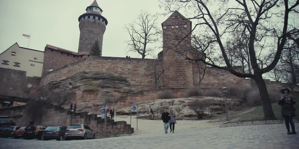 People walk along a cobblestone path toward a historic stone castle surrounded by bare trees on an overcast day