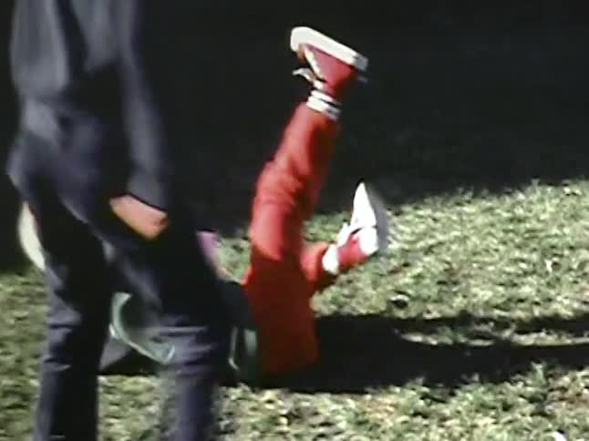 A young boy wearing a football helmet and red pants runs across a grassy backyard before falling down during a game with other children