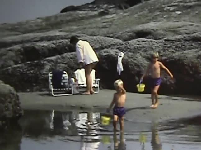 Two young boys on a beach carrying yellow buckets and playing around while waving to the camera with a young adult woman on the background sitting on a chair outdoors during day time