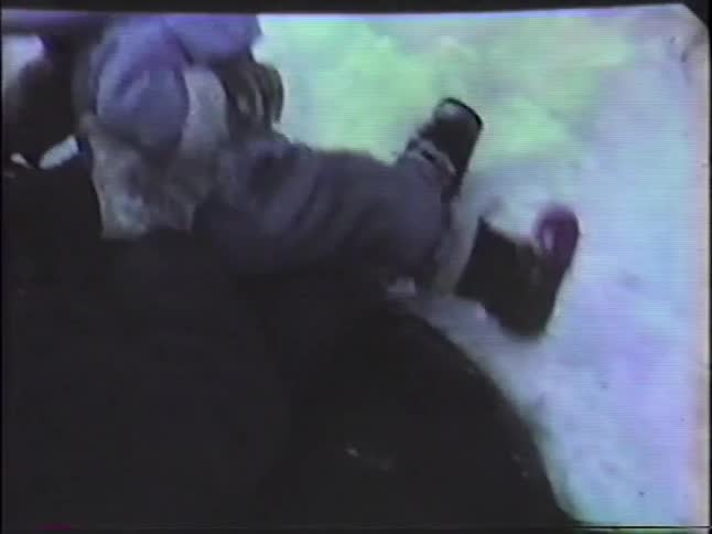 A young child in a purple snowsuit and pompom hat laughs while sledding down a snowy hill with an adult