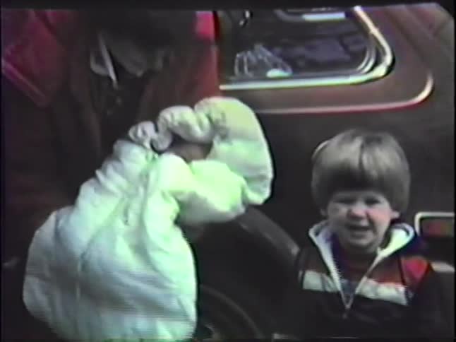 Young adult woman holding a baby wrapper in a blanket with a toddler boy next to her and a car and trees on the background outdoors during day time