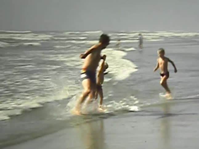In this vintage video a man holds a small childs hand to help them balance on a skimboard as it glides across the shallow wateras the board slows down and the child hops off another young boy is seen running nearby on the foggy shoreline