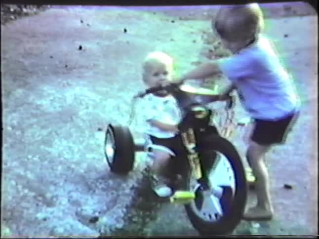 In a grainy faded home video a young boy carefully pushes his toddler brother who is sitting on a large plastic tricycle