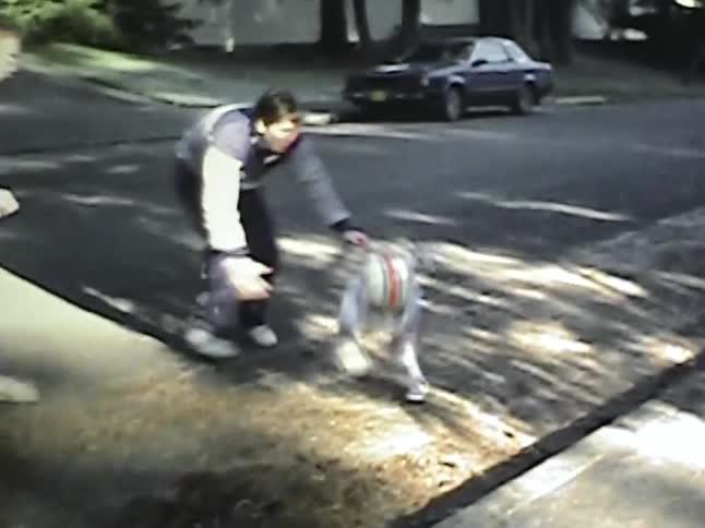 A young child wearing a football helmet and jersey runs across a street while being playfully chased by an adultthe child trips and falls near the curb and two adults quickly step in to help them back up to their feet