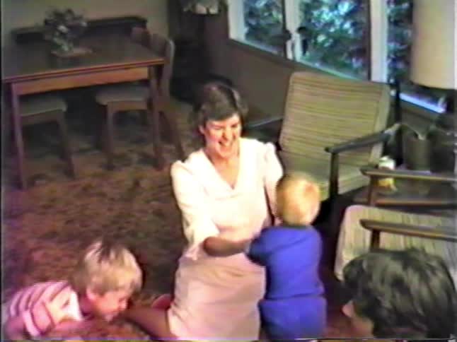 A woman kneels on a carpeted floor and smiles while guiding a toddler in a blue outfit as he takes small steps toward her while another young child moves around them in a sunlit living room featuring vintage furniture and large windows