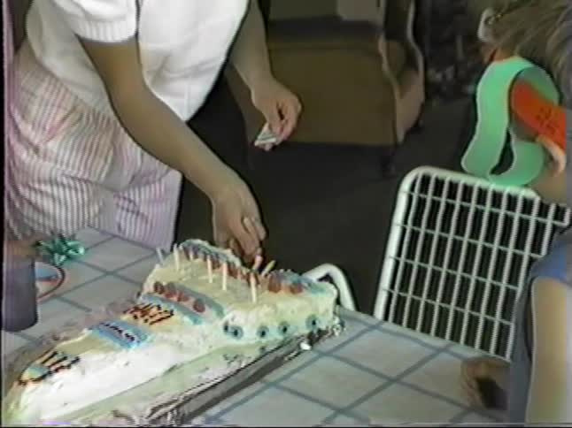 Mother lights the birthday candles while children gather around a cake on the dining table at home