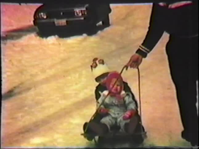 A man walks through a snowy street at night pulling two young children bundled in winter gear on a small sledthe scene captures a nostalgic homemovie aesthetic as the group passes parked cars under the glow of streetlights