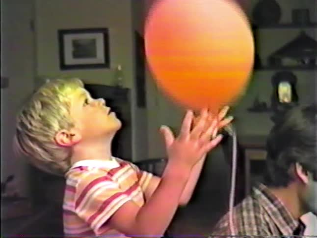 A young boy playing with a red balloon on a string while his family sits nearby