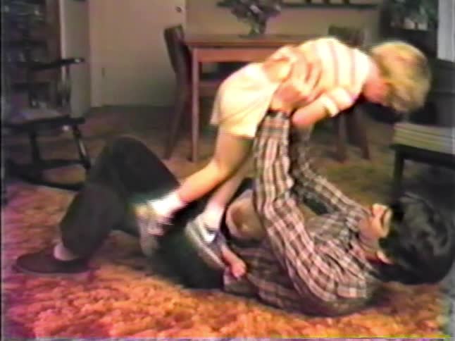 Young adult man lying on the floor with a toddler girl over him and a boy coming and playfully struggling to bend his arm before falling to the floor in a living room indoors during day time