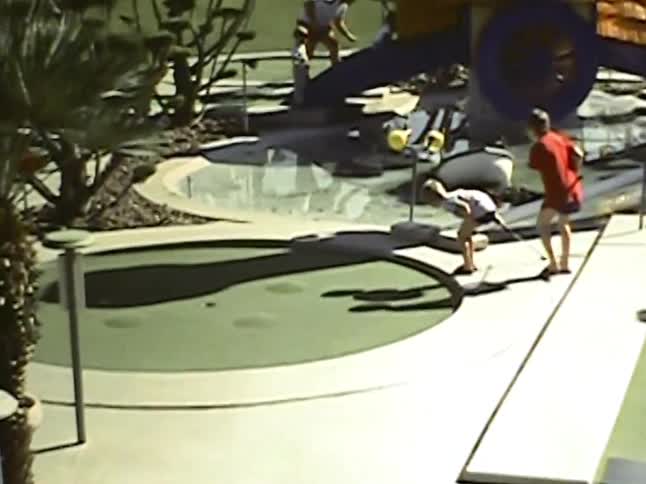 In grainy vintage footage from a sunny day a young boy in a red shirt putts on a minigolf course while his family watches the ball roll toward the next hole
