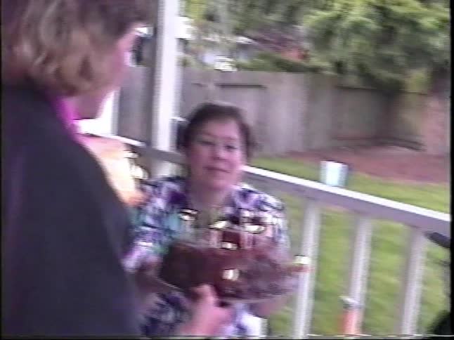 A woman carries two chocolate and vanilla cakes with lit candles out to a wooden deck where a woman and children wait at a table with a red checkered cloth to celebrate a birthday