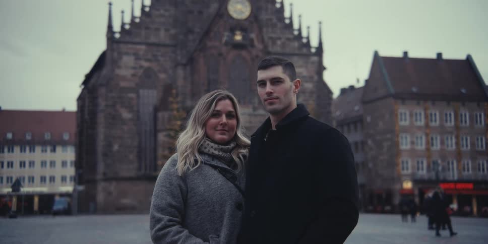 A couple stands for a solemn portrait in a historic city square their serious expressions framed by the grand facade of an ancient church behind them