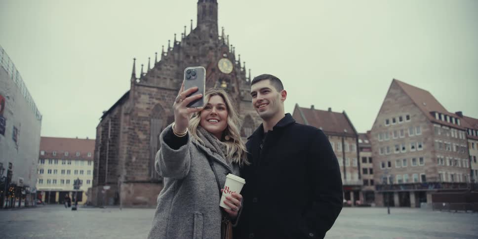 A happy couple stops while exploring a historic town square the woman holding up her phone to capture a smiling selfie together as her partner kisses her on the cheek and then they share a kiss
