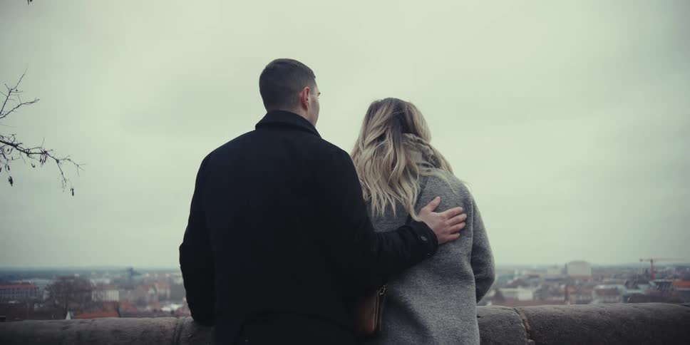 Man and woman standing together with his arm around her and he kisses her as they stand near a railing overlooking neighborhood buildings below
