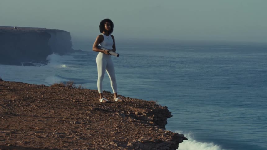 Woman standing on a seaside cliff holding a training mat with the ocean in the background