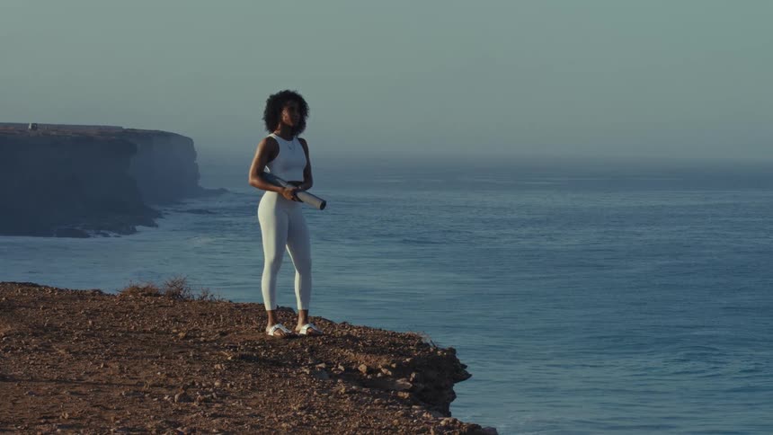 Woman stands on a cliff overlooking the ocean while holding a cylindrical object