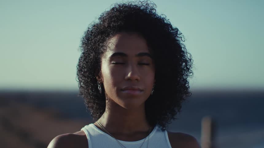 A young woman stands with eyes closed and taking a calm deep breath in natural light outdoors
