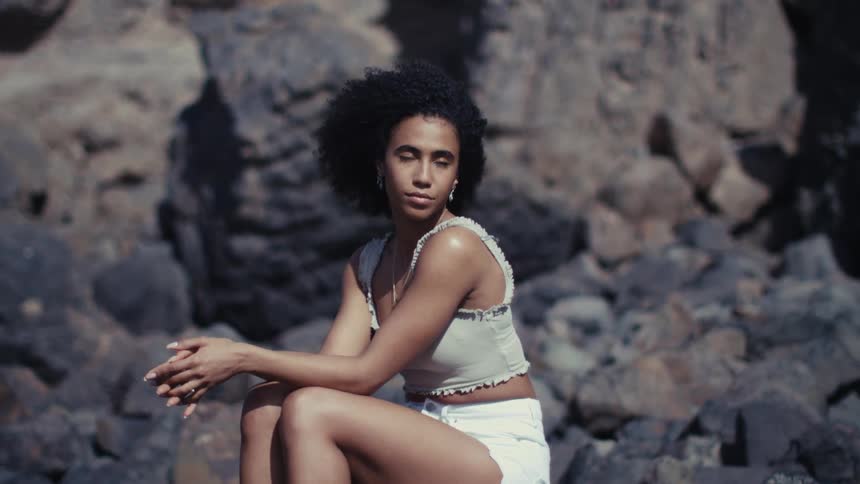 A woman with curly hair sits on dark rocks against a rugged cliffside under bright sunlightshe wears a lightcolored tank top and white shorts looking thoughtfully into the distance while adjusting her hair