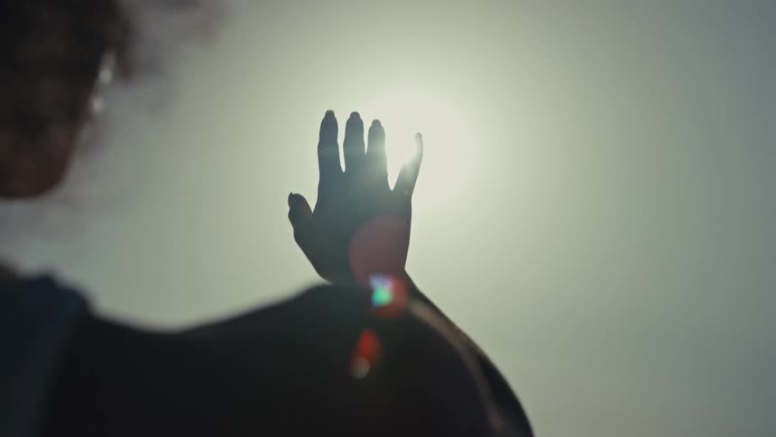 A young woman outdoors lifts her right hand to shield her eyes using it to block the warm light of the setting sun