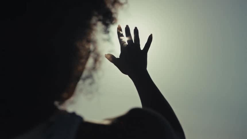 Young adult black woman standing and reaching her hand out towards the sun on the sky outdoors during day time