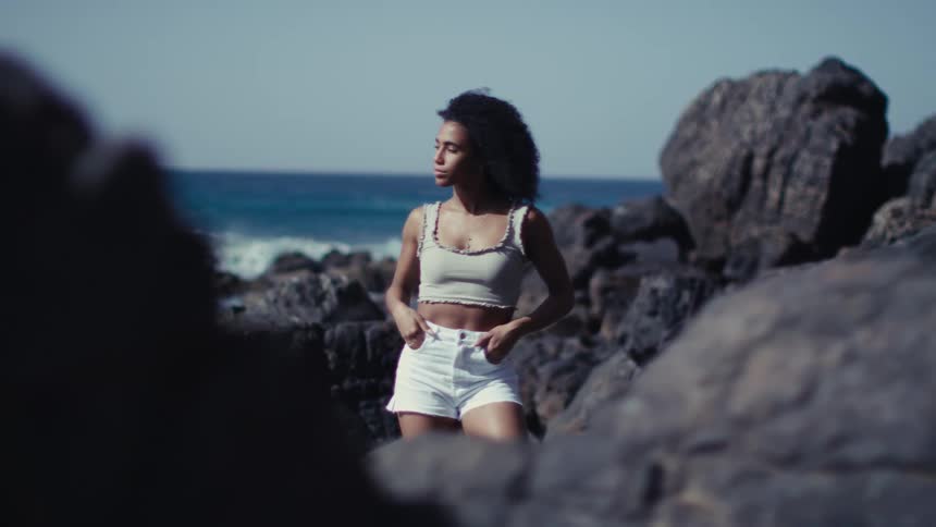 Woman wearing a summer outfit stands among the boulders on the shore on a windy summer day