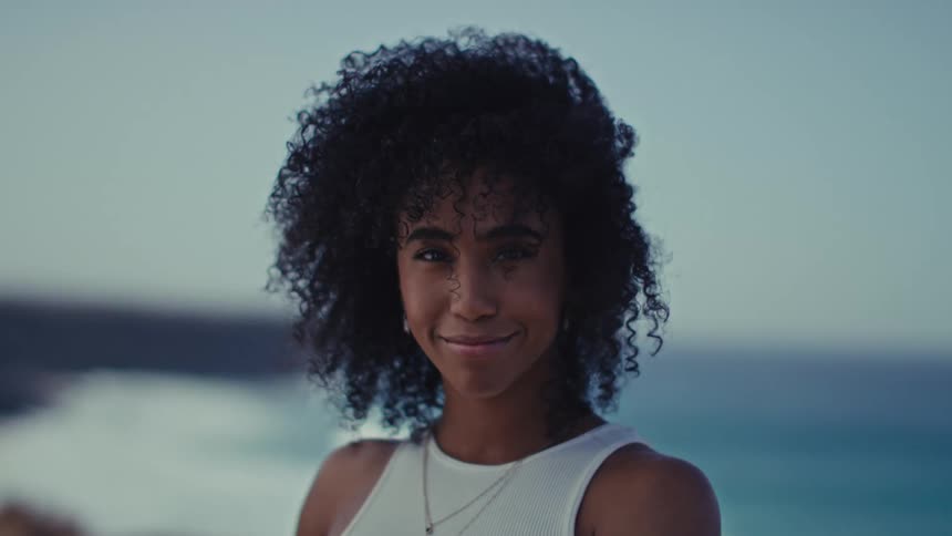 Against a softfocus ocean backdrop a woman with voluminous curly hair looks into the camera her gentle expression blooming into a bright warm smile