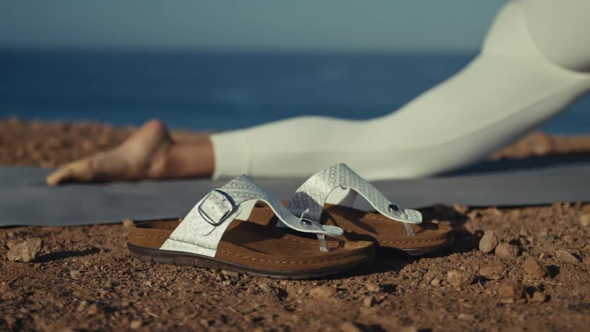 A pair of white sandals sits on the rocky ground in the foreground while a person practices yoga on a mat in the background the scene is set outdoors on a cliff overlooking the blue ocean under bright daylight