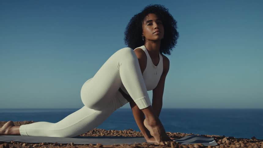 A woman in white athletic wear performs a deep lunge and a complex yoga bind on a mat overlooking the oceanshe looks directly toward the camera before lowering her gaze to focus on her posture and balance against the clear blue sky