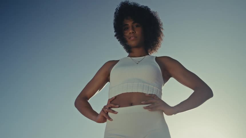 A woman with dark curly hair stands confidently against a bright clear sky she wears a white athletic twopiece outfit and rests her hands on her hips while looking directly at the camera