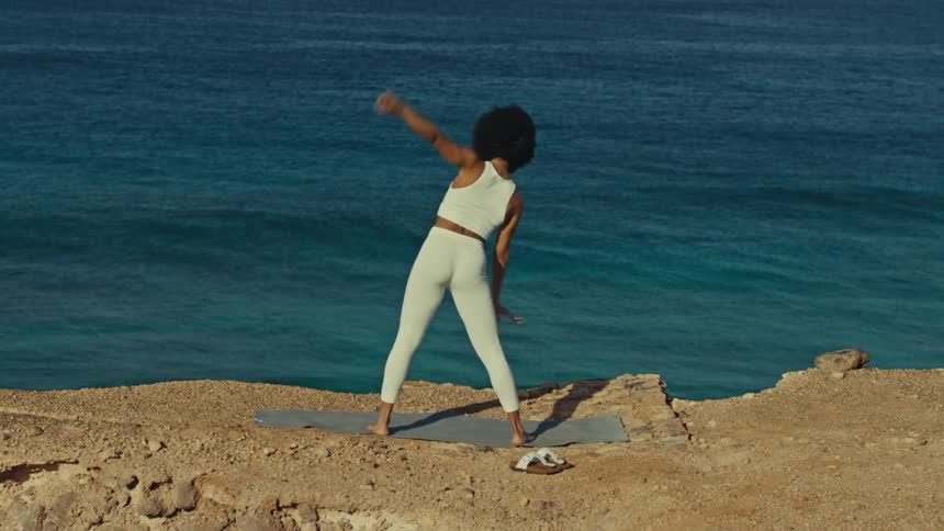 Woman stretches while standing on a yoga mat at the shore on a summer day