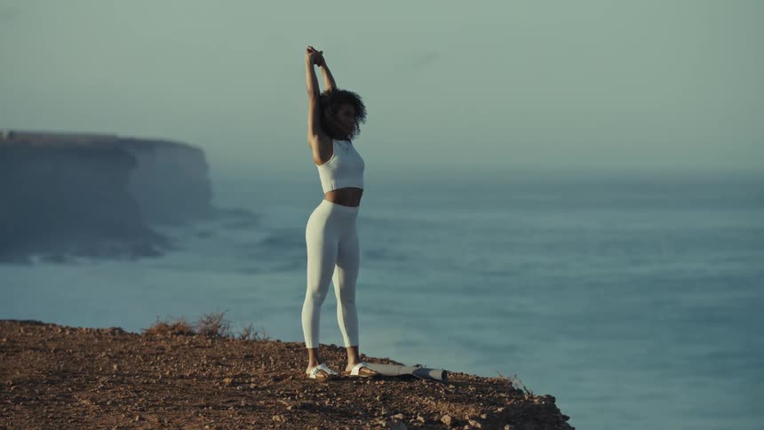 A young woman wearing yoga tights stands on a cliff at midday stretching as sunlight highlights the landscape around her