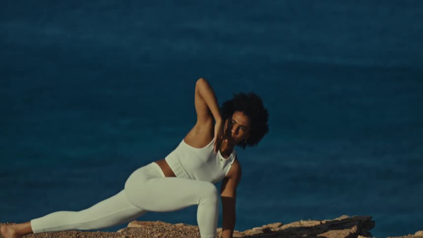 A woman stretches in a yoga pose on a rocky cliff beside the ocean at sunrise