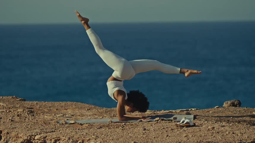 Woman performs a yoga inversion on a mat atop a cliff with the ocean in the background