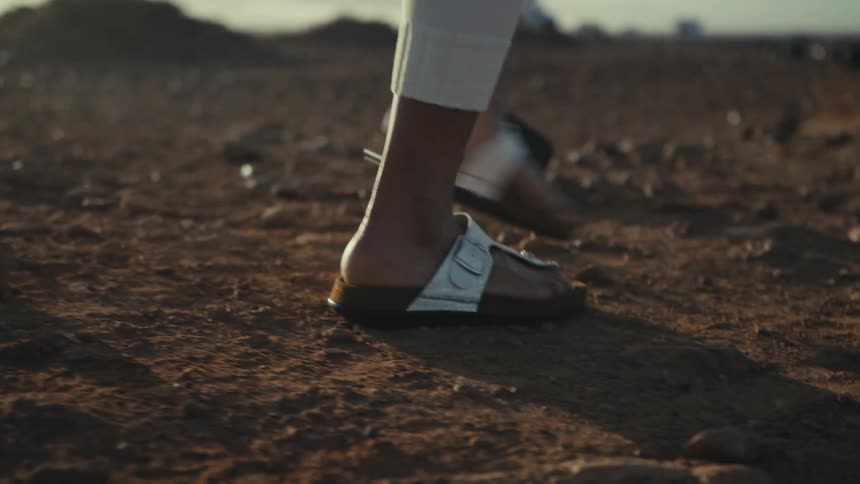 Feet of a black female person wearing sandals and white pants walking towards a cliff next to the ocean outdoors during dusk or dawn