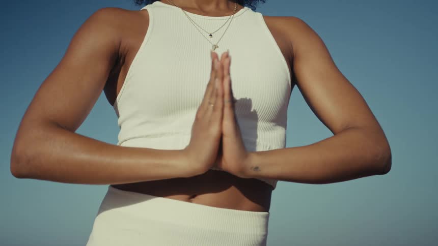 A young woman with curly hair is shown outdoors against a clear blue sky wearing a white athletic top she begins by adjusting her hair and then brings her hands together in a prayer position at her chest appearing focused and serene