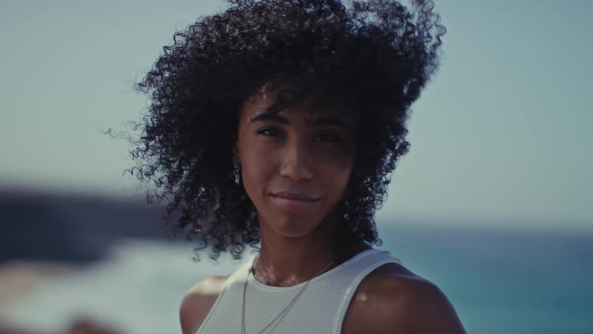 With her curly hair gently blowing in the wind a confident woman looks directly into the camera and offers a soft warm smile against a serene ocean backdrop