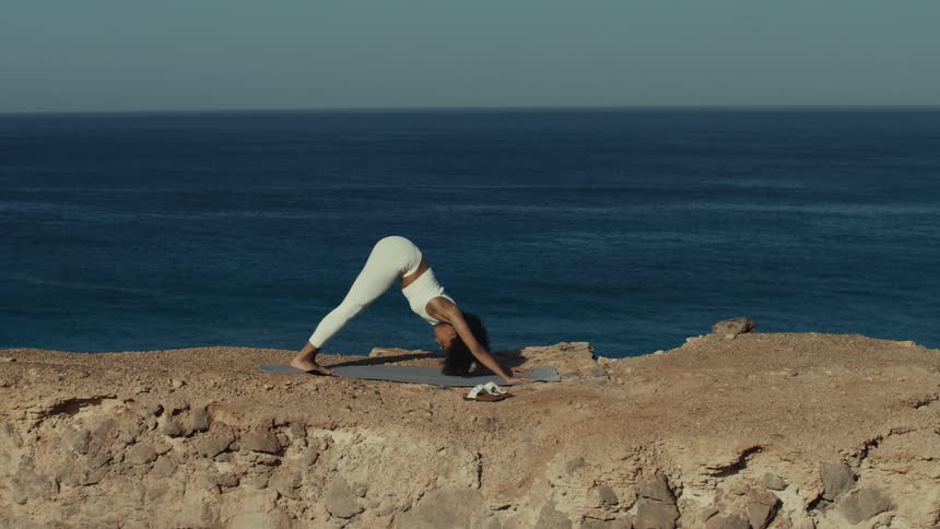 Woman performing a stretching pose on a training mat atop a cliff with the ocean in the background