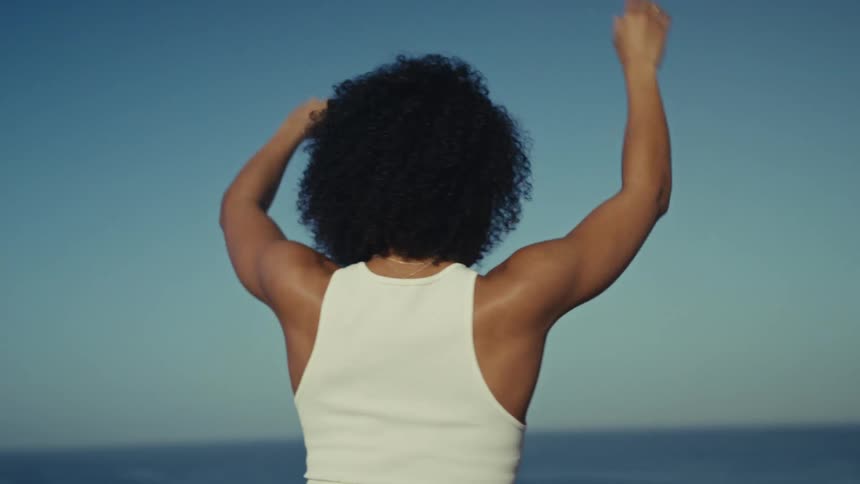 Woman raises her arms above her head with the sky and sea in the background
